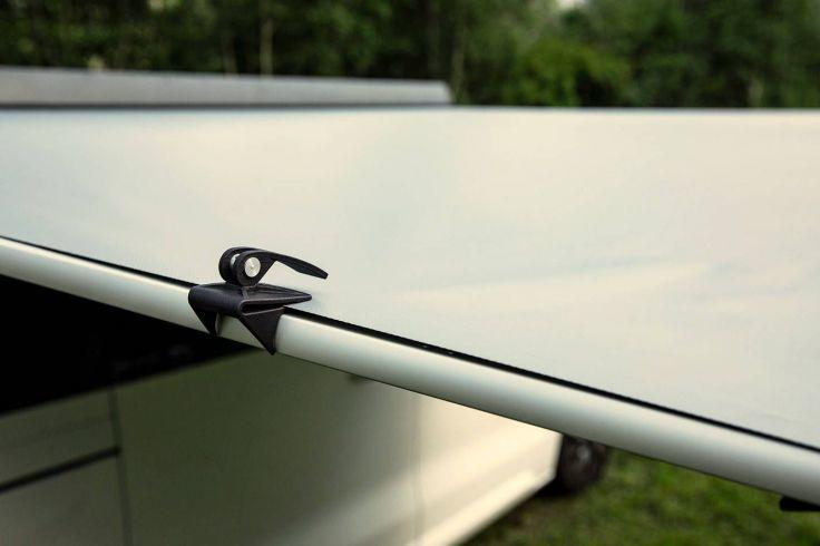 Nahaufnahme einer ausgefahrenen Markise an der Seite eines weißen Fahrzeugs, sichtbare Metallklemme, Gras und Bäume im Hintergrund