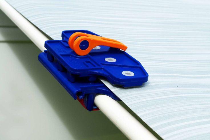 Blaue Kunststoffklemme mit orangefarbenem Hebel an der Kante einer durchsichtigen, geriffelten Kunststoffplatte neben einem weißen Rundrohr