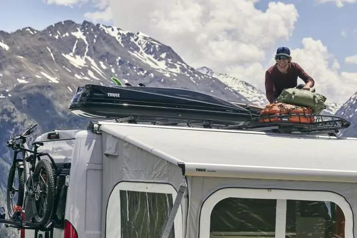Wohnmobil mit Dachbox und Gep&auml;ck, eine Person auf dem Dach, zwei Fahrr&auml;der an der R&uuml;ckseite, schneebedeckte Berge im Hintergrund und bew&ouml;lkter Himmel