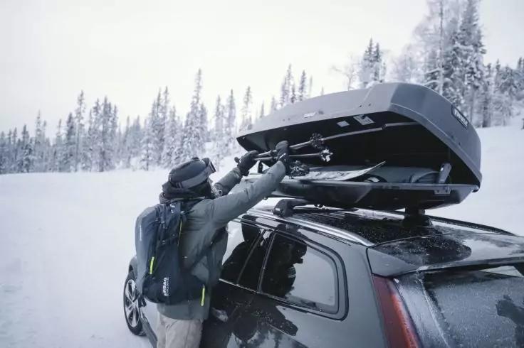 Person in Winterkleidung verstaut lange Ausr&uuml;stung in ge&ouml;ffnetem Dachkoffer auf einem dunklen Auto; verschneite Landschaft mit Nadelb&auml;umen im Hintergrund