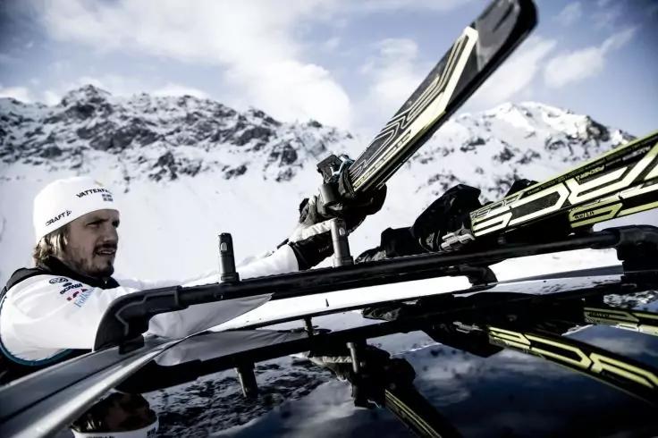 Person mit wei&szlig;er M&uuml;tze setzt Skier auf den Dachtr&auml;ger eines Autos, im Hintergrund schneebedeckte Berge und Himmel