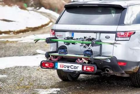 Heck eines silbernen SUV mit montiertem Hecktr&auml;ger, darauf zwei Skier und zwei Snowboards; Boden mit Schneeresten und Kies