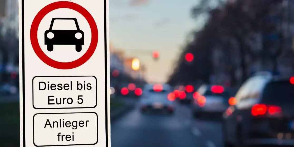 Verkehrsschild links: roter Kreis mit Autosymbol und die Aufschriften 'Diesel bis Euro 5' und 'Anlieger frei'; verschwommene Autos mit Bremslichtern auf einer Stra&szlig;e im Hintergrund
