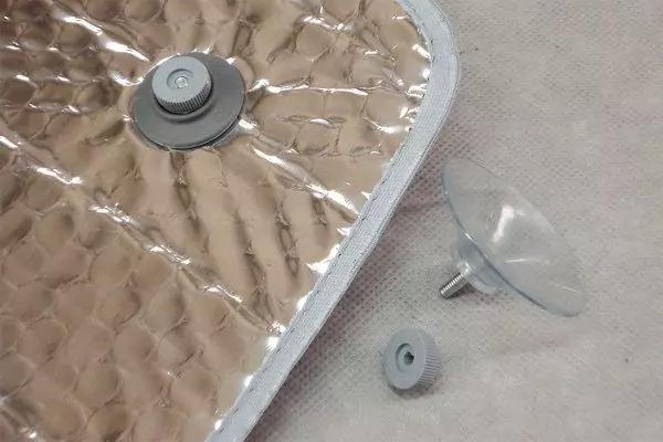 Silberne, reflektierende rechteckige Matte mit gepr&auml;gtem Wabenmuster, ein grauer runder Kunststoffknopf, eine lose graue Kunststoffkappe und ein durchsichtiger Saugnapf mit Metallstift auf hellem Stoff