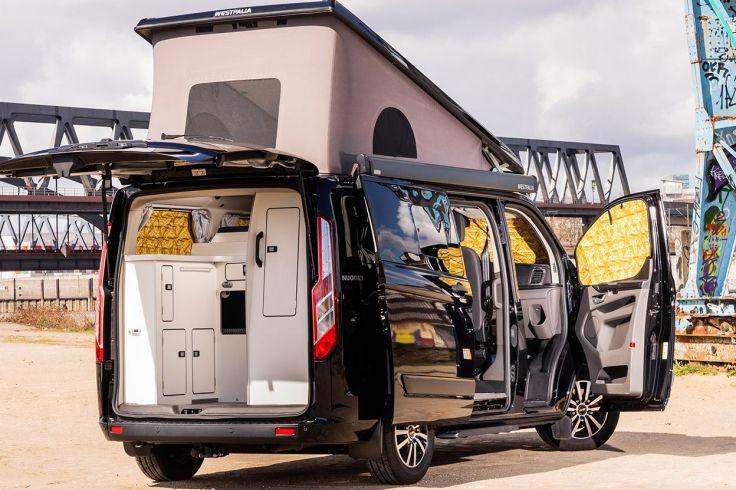 Schwarzer Campingbus mit ge&ouml;ffneten Heck- und Seitent&uuml;ren, sichtbare wei&szlig;e K&uuml;chenschr&auml;nke und gelbe Vorh&auml;nge, im Hintergrund Br&uuml;ckenbauwerk und Graffiti