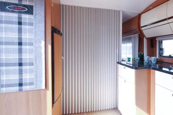 Innenraum mit geschlossener Faltwand/Trennvorhang, links kariertes Vorhang, K&uuml;hlschrank, helle Holzschr&auml;nke, schwarze Arbeitsplatte mit Sp&uuml;le und kleinen Flaschen