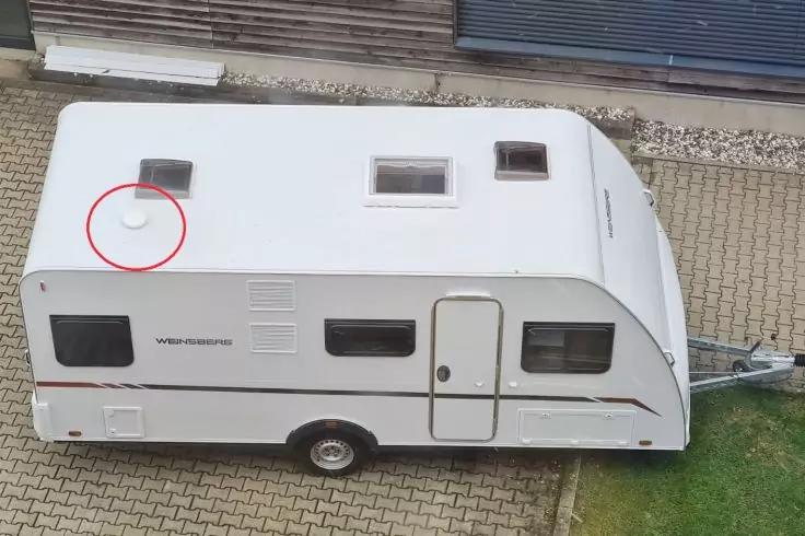 Weißer Wohnwagen auf gepflastertem Stellplatz, Seitenansicht mit Tür, Fenstern, einem Rad, zwei Dachluken und einer runden weißen Scheibe auf dem Dach, die mit einem roten Kreis markiert ist; rechts ein Grasstreifen