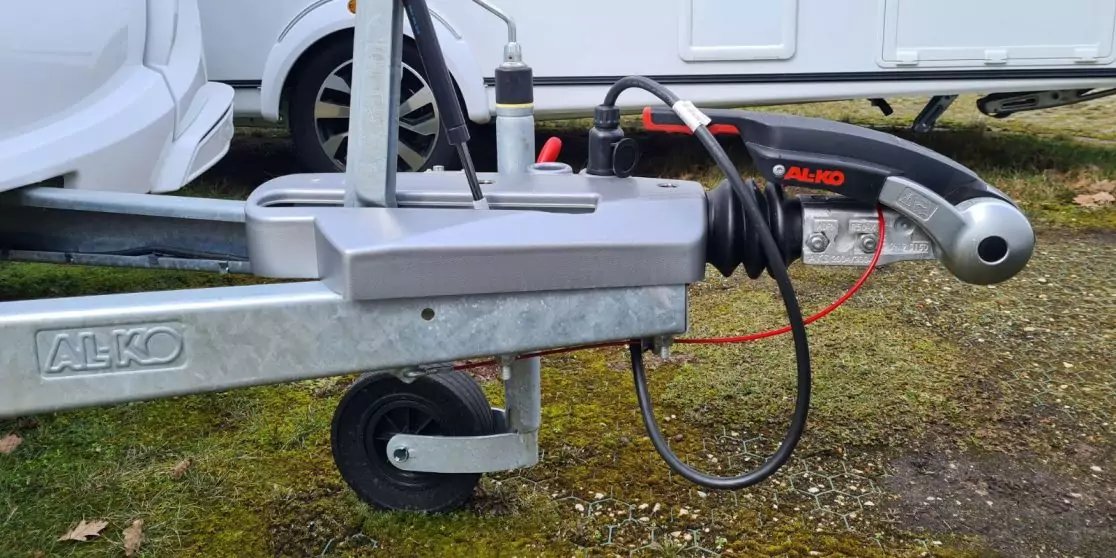 Anh&auml;nger-Kupplung mit Metall-Dissel und AL-KO-Schriftzug, Kupplungskopf, aufgerollenes schwarzes Kabel, rotes Kabel, St&uuml;tzrad, wei&szlig;es Fahrzeug im Hintergrund, Gras- und Schotterboden