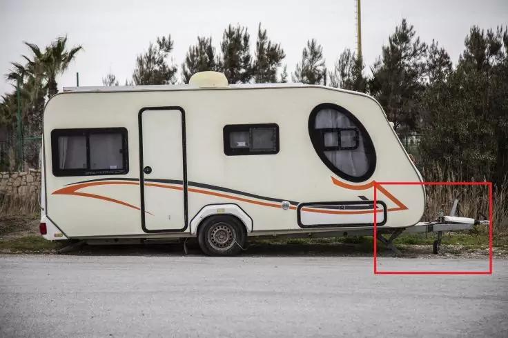 Wei&szlig;er Wohnwagen mit orangefarbenen Streifen, zwei rechteckige Fenster und ein rundes Frontfenster, an der Stra&szlig;e geparkt; Zugstange vorne im roten Rahmen; B&auml;ume im Hintergrund