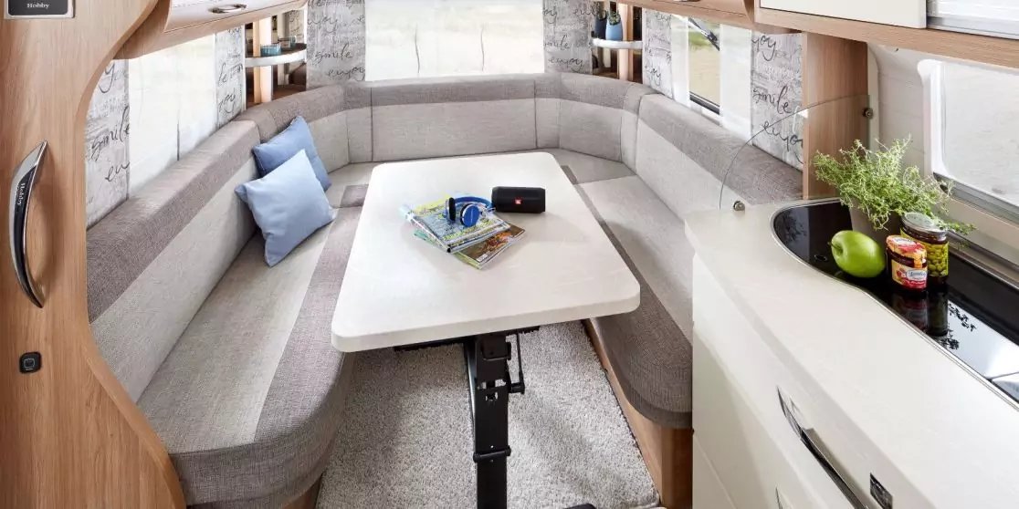 Innenraum eines Wohnmobils mit halbrundem Sitzbereich mit grauen Polstern, mittigem Tisch, K&uuml;chenzeile mit Sp&uuml;le, &Auml;pfeln und Kr&auml;utern auf der Arbeitsplatte, Fenstern mit Vorh&auml;ngen