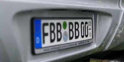 Nahaufnahme des Fahrzeughecks mit wei&szlig;em deutschen Kennzeichen mit schwarzen Zeichen FBB BB 00, blauem EU-Streifen mit D und zwei runden Aufklebern