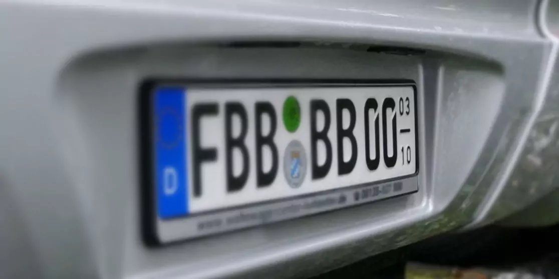 Nahaufnahme des Fahrzeughecks mit wei&szlig;em deutschen Kennzeichen mit schwarzen Zeichen FBB BB 00, blauem EU-Streifen mit D und zwei runden Aufklebern