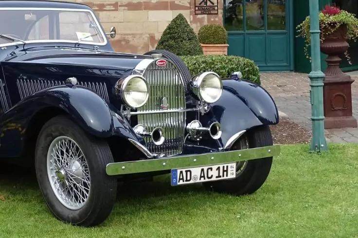 Vorderer Bereich eines dunkelblauen Autos mit chromfarbenem K&uuml;hlergrill, zwei runden Scheinwerfern, Sto&szlig;stange und Kennzeichen "AD-AC 1H". Vorderes linkes Drahtspeichenrad sichtbar. Auto steht auf Rasen vor einem Geb&auml;ude mit gr&uuml;ner T&uuml;r, Topfpflanzen