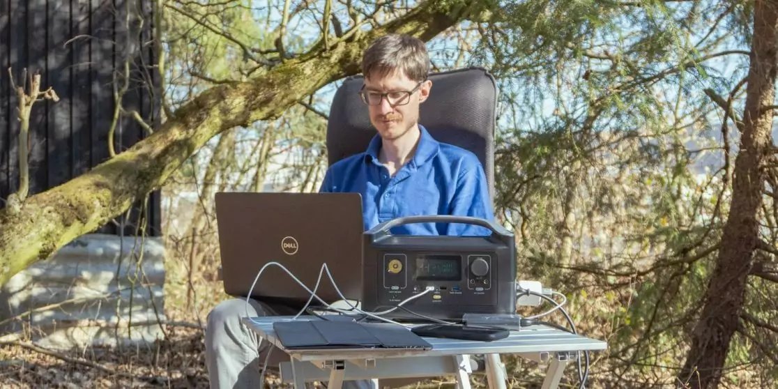 Person mit Brille und blauem Hemd sitzt drau&szlig;en auf einem Stuhl an einem Klapptisch mit Laptop und tragbarer Stromstation; Kabel auf dem Tisch, B&auml;ume und Holzh&uuml;tte im Hintergrund