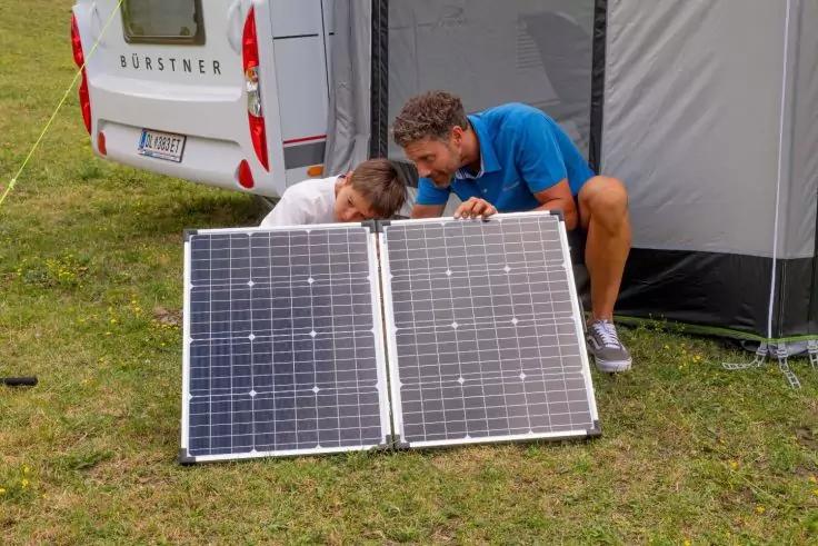 Zwei Personen hocken neben einem ausgeklappten, faltbaren Solarpanel auf einer Grasfl&auml;che; im Hintergrund ein Wohnmobil und ein Vorzelt