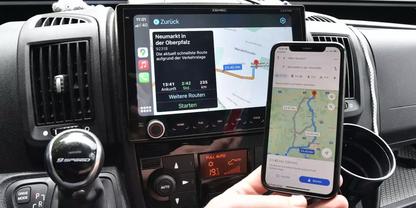Autoarmaturenbrett mit mittigem Touchscreen, der eine Kartenroute zeigt; eine Hand h&auml;lt ein Smartphone mit ebenfalls sichtbarer Kartenroute; Schalthebel, Luftausl&auml;sse und Bedienelemente sichtbar