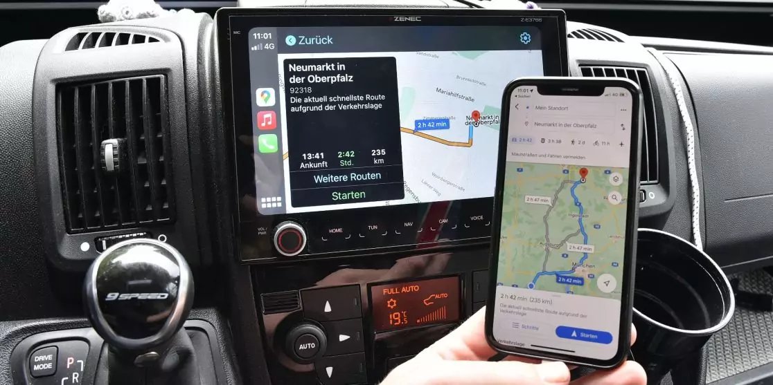 Autoarmaturenbrett mit mittigem Touchscreen, der eine Kartenroute zeigt; eine Hand h&auml;lt ein Smartphone mit ebenfalls sichtbarer Kartenroute; Schalthebel, Luftausl&auml;sse und Bedienelemente sichtbar