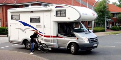 Wei&szlig;es Wohnmobil auf einer Stra&szlig;e, davor eine stehende Person mit Fahrrad, H&auml;user und B&auml;ume im Hintergrund