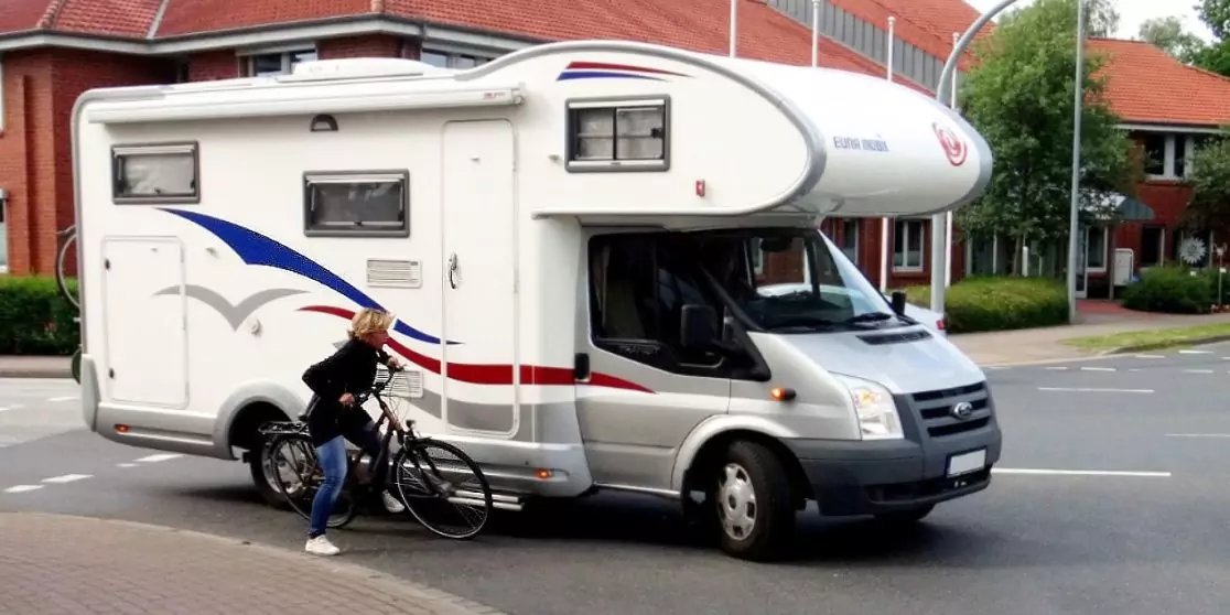 Wei&szlig;es Wohnmobil auf einer Stra&szlig;e, davor eine stehende Person mit Fahrrad, H&auml;user und B&auml;ume im Hintergrund