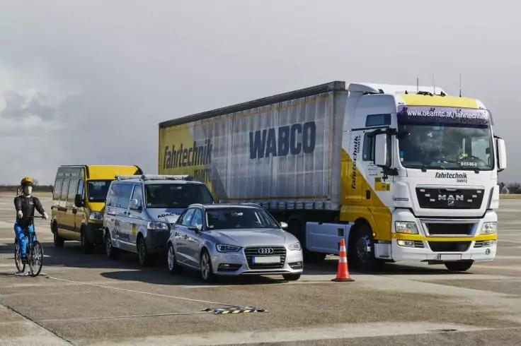 Lkw mit Anh&auml;nger, silberner Pkw, gelber Kleinbus, Transporter, eine Person mit Fahrrad, ein orangefarbener Verkehrskegel auf einer Asphaltfl&auml;che unter bew&ouml;lktem Himmel