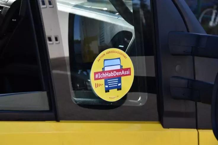 Runder gelber Aufkleber mit rotem Rechteck und wei&szlig;em Text '#IchHabDenAssi' an der Innenseite einer Seitenscheibe eines gelben Fahrzeugs; sichtbarer T&uuml;rgriff und Au&szlig;enspiegel
