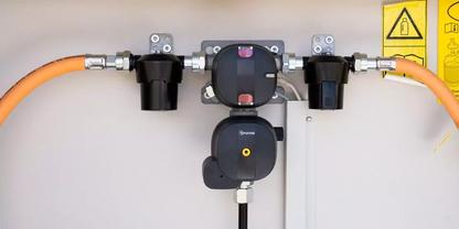 Mehrere schwarze, runde Anschlussbauteile an einer grauen Wand, verbunden mit orangefarbenen Kabeln; rechts oben ein gelbes Warnsymbol