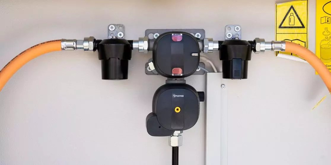 Mehrere schwarze, runde Anschlussbauteile an einer grauen Wand, verbunden mit orangefarbenen Kabeln; rechts oben ein gelbes Warnsymbol