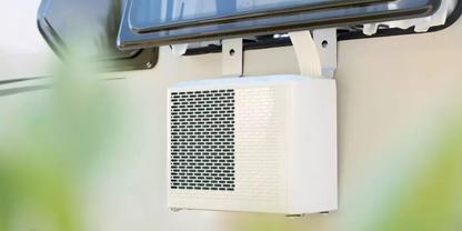 Wei&szlig;es rechteckiges Ger&auml;t mit gelochter Frontplatte, an einer hellen Au&szlig;enwand unter einem ge&ouml;ffneten Fenster montiert; zwei Metallhalterungen sichtbar; unscharfe gr&uuml;ne Pflanzen im Vordergrund