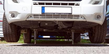 Tiefenperspektive der Front eines wei&szlig;en Autos, sichtbare Unterseite, zwei Metallst&uuml;tzen unter dem Wagen, Vorderr&auml;der, Nummernschild, Kiesboden