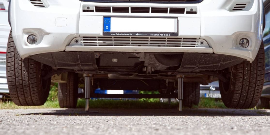 Tiefenperspektive der Front eines wei&szlig;en Autos, sichtbare Unterseite, zwei Metallst&uuml;tzen unter dem Wagen, Vorderr&auml;der, Nummernschild, Kiesboden