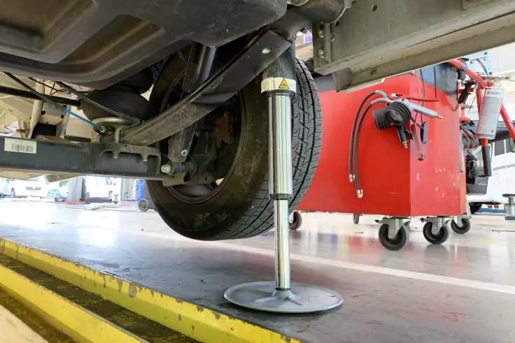 Unterseite eines Fahrzeugs mit sichtbarem Rad und Achse, ausgeklappte Metallst&uuml;tze mit rundem Fu&szlig; auf Werkstattboden, roter Werkzeugwagen mit Rollen im Hintergrund, gelbe Bodenmarkierung