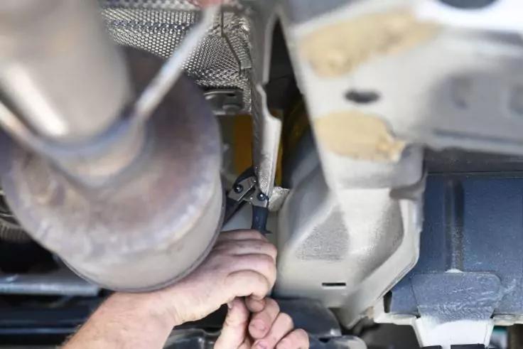 Zwei H&auml;nde halten einen Schraubenschl&uuml;ssel unter einem Auto; sichtbarer Auspuff, Halterung und Unterboden