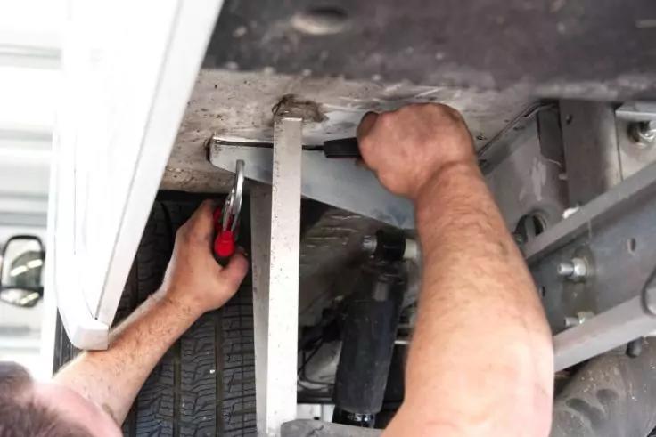 Arme einer Person unter einem Fahrzeug; rechte Hand h&auml;lt ein Metallteil, linke Hand h&auml;lt eine Zange mit rotem Griff; sichtbarer Reifen und Fahrzeugrahmen