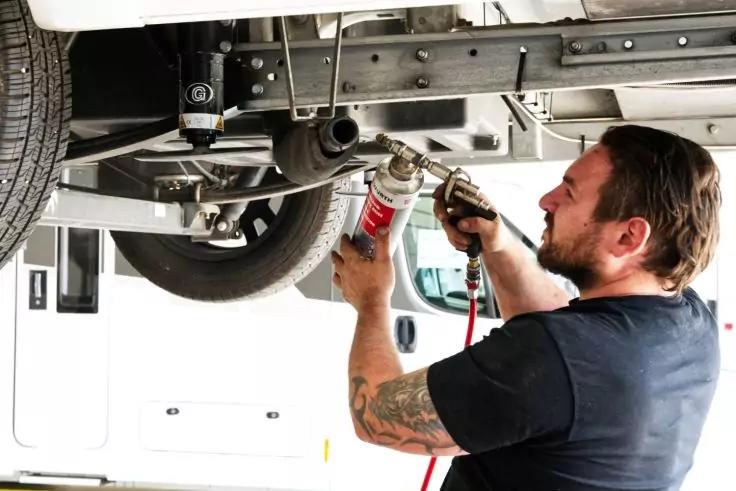 Person mit schwarzem T‑Shirt und T&auml;towierung am Unterarm benutzt ein pneumatisches Werkzeug an der Unterseite eines Fahrzeugs; Rad und Fahrgestell sichtbar