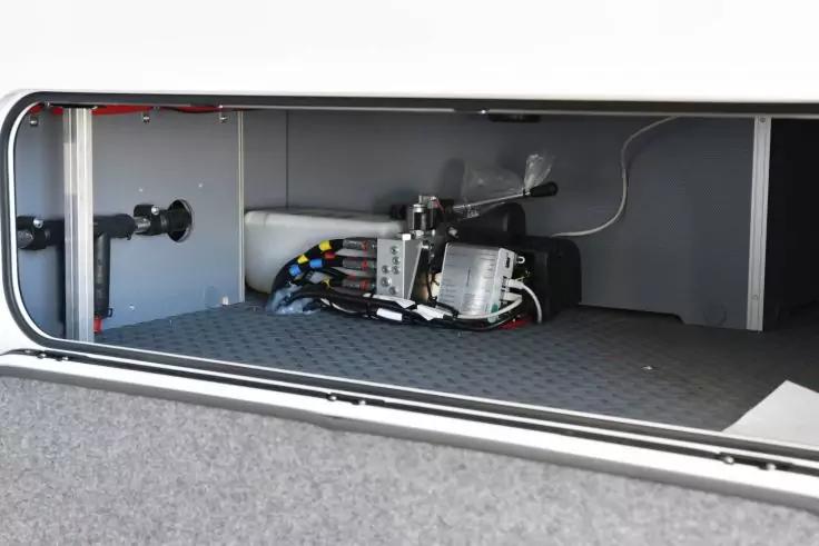 Offenes Abteil mit grauem, strukturiertem Boden; B&uuml;ndel farbiger Kabel und zwei silberne Metallgeh&auml;use mit angeschlossenen Leitungen; wei&szlig;e Innenw&auml;nde und Rahmen