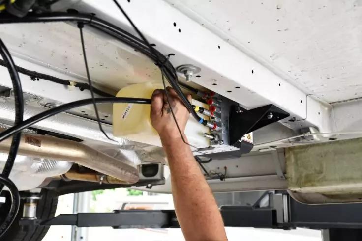 Unterseite eines Fahrzeugs mit Kabeln, Steckverbindern und einer gelben Plastikflasche; eine Hand h&auml;lt die Anschl&uuml;sse