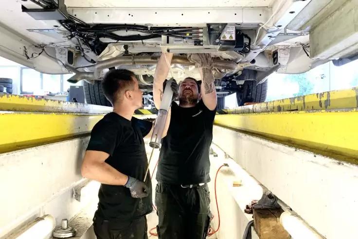 Zwei Personen in einer Grube unter einem angehobenen Auto; eine h&auml;lt ein Werkzeug &uuml;ber Kopf