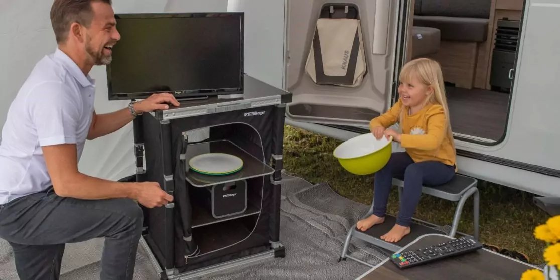 Mann kniet neben offenem schwarzem mobilen Schrank mit gestapelten Tellern; blondes M&auml;dchen sitzt auf Klappstuhl und h&auml;lt eine wei&szlig;e Sch&uuml;ssel; Wohnmobil im Hintergrund