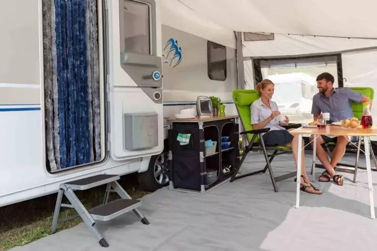 Zwei Personen sitzen an einem Campingtisch unter dem Vorzelt eines Wohnmobils; grüne Klappstühle, Tisch mit Getränken und Snacks, Regal mit Utensilien, geöffnete Wohnmobiltür, Bodenmatte