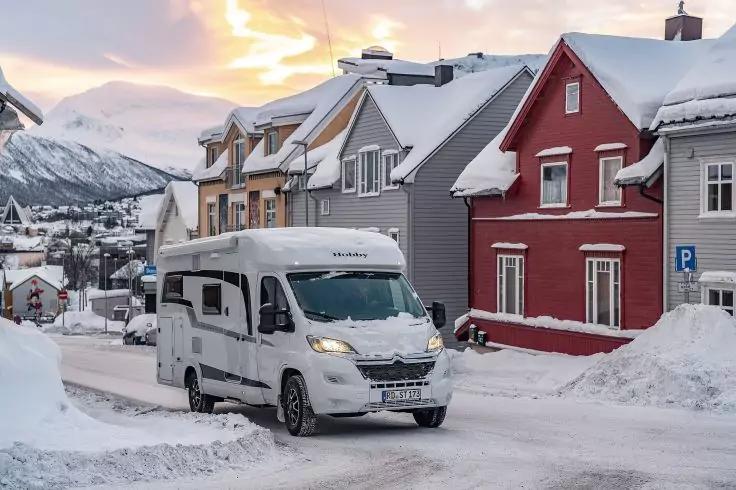 Wohnmobil auf verschneiter Stra&szlig;e vor bunten H&auml;usern mit schneebedeckten D&auml;chern und Bergen im Hintergrund, heller Himmel