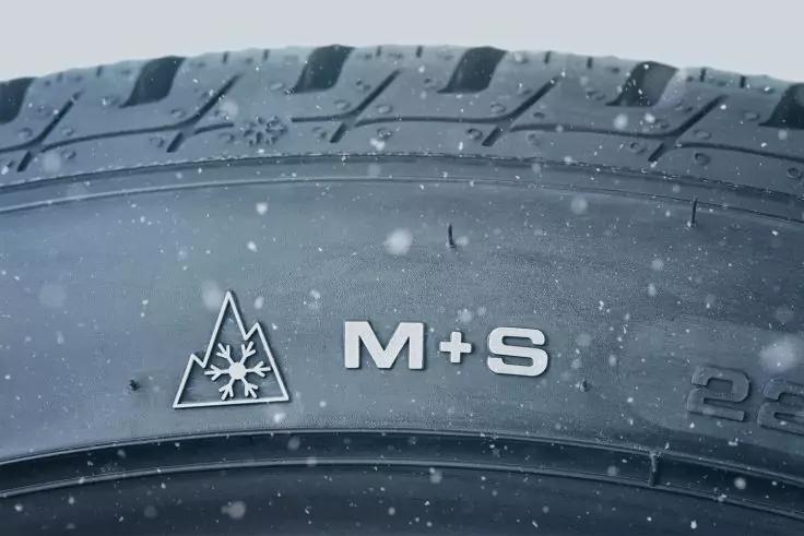Nahaufnahme einer Reifenflanke mit erhabenen Buchstaben "M+S" und einem Bergsymbol mit Schneeflocke, kleine Schneepartikel auf dem Reifen