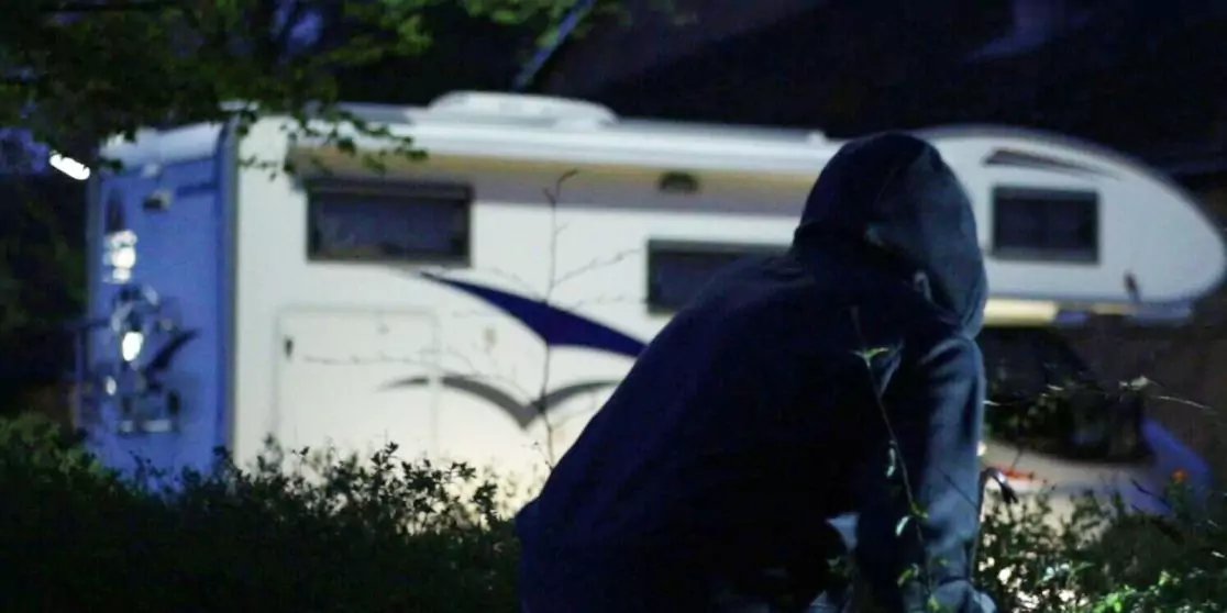 Person in dunkler Kapuzenjacke beugt sich nachts an einem Strauch, wei&szlig;es Wohnmobil im Hintergrund