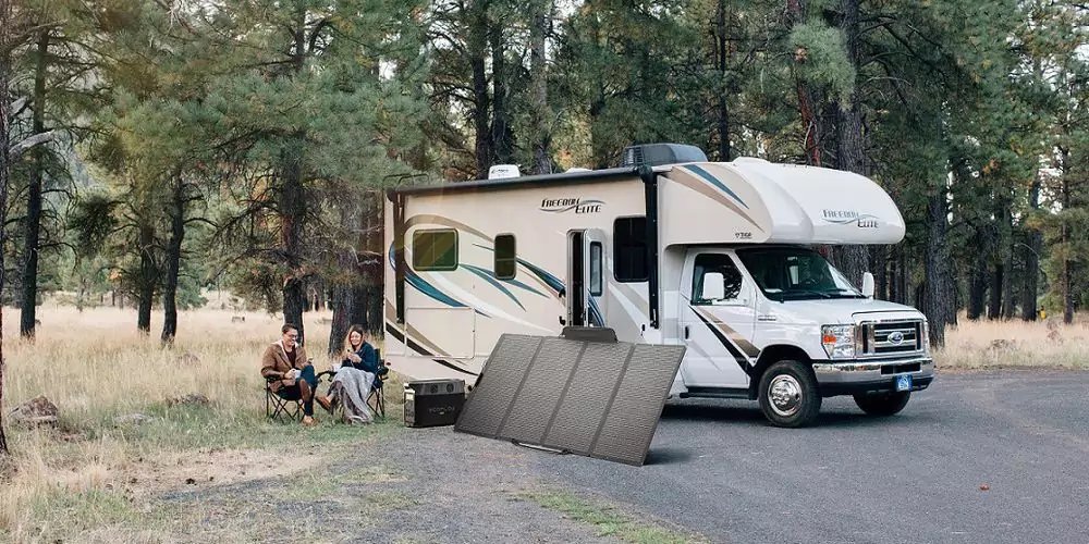 Wohnmobil geparkt an einem Wald-Campingplatz; zwei Personen sitzen auf Campingst&uuml;hlen neben dem Fahrzeug; aufgeklappte Solarpaneele liegen auf dem Boden vor dem Wohnmobil; Nadelb&auml;ume und Gras im Hintergrund