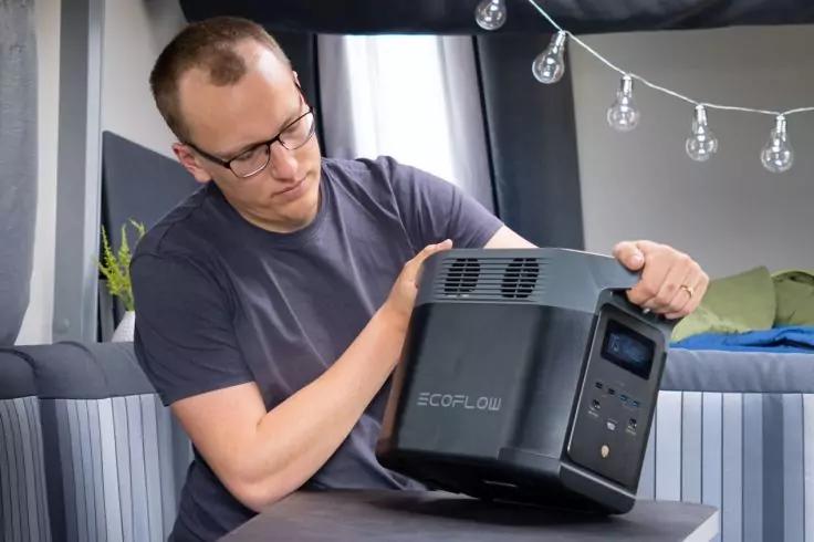 Person mit Brille h&auml;lt ein schwarzes, quaderf&ouml;rmiges Ger&auml;t mit Display, L&uuml;ftungsgittern und 'ECOFLOW'-Schriftzug in einem Raum mit Sofa und Lichterkette