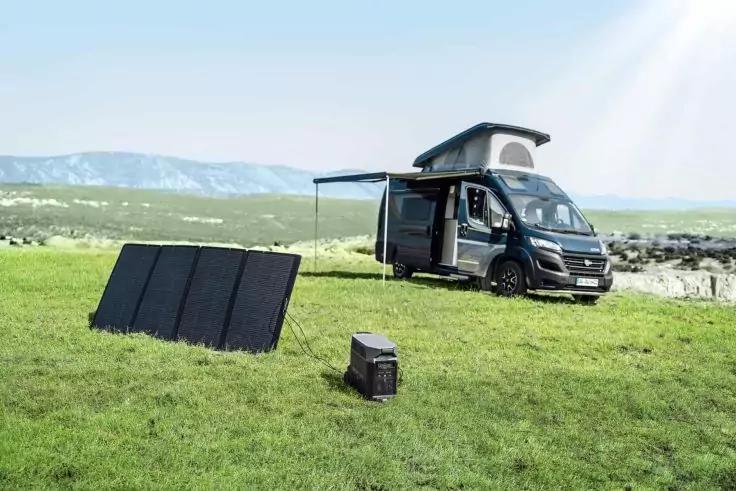 Schwarzes Wohnmobil mit ge&ouml;ffnetem Aufstelldach und ausgezogenem Sonnensegel auf gr&uuml;ner Wiese; davor Solarpanels auf St&auml;nder und tragbare Batterie; Felsen und Himmel im Hintergrund