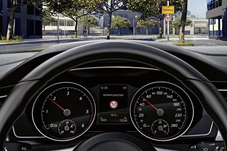Blick &uuml;ber Lenkrad auf Instrumententafel: links Drehzahlmesser, rechts Tachometer, mittiges Display zeigt Verkehrsschild '50'; durch die Windschutzscheibe Stra&szlig;e mit B&auml;umen und Geb&auml;uden