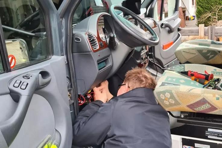 Eine Person mit Oberk&ouml;rper unter dem Armaturenbrett eines Fahrzeugs; Lenkrad, offene T&uuml;r, Werkzeuge und Sitzpolster sichtbar