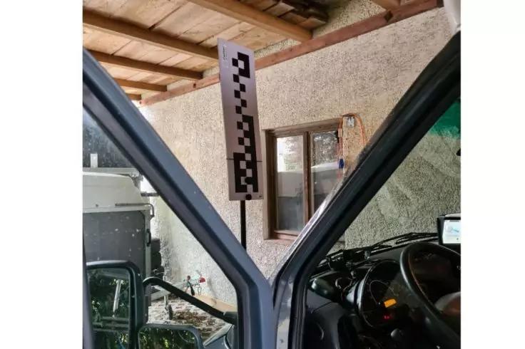 Ge&ouml;ffnete vordere Autot&uuml;ren, Lenkrad und Armaturenbrett sichtbar, mittig vertikale Tafel mit schwarz‑wei&szlig;em Schachbrettmuster, dahinter Wand mit kleinem Fenster und Holzdecke, links Fahrradrad sichtbar