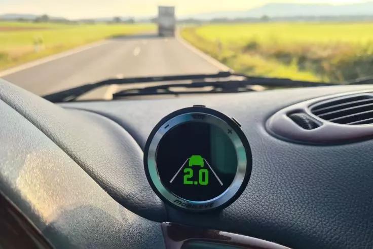 Rundes digitales Display auf dem Armaturenbrett mit gr&uuml;nem Auto-Symbol und der Zahl "2.0", teilweise sichtbares Lenkrad, gerade Stra&szlig;e mit einem Lkw in der Ferne, Felder an beiden Seiten und heller Himmel