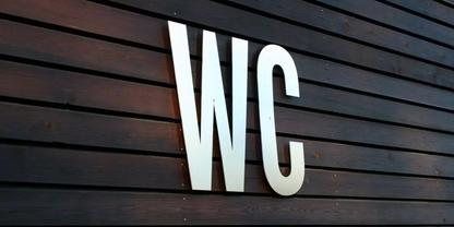Wei&szlig;e Gro&szlig;buchstaben 'WC' auf dunkelbraunen, horizontalen Holzlatten